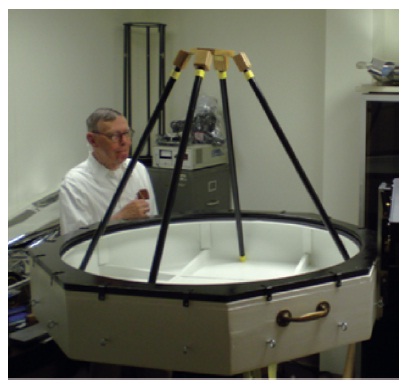 Koch is observing the wooden mirror cell constructed by a carpentry student from a local trade school. Variable length truss tubes are temporarily setting inside the mirror cell. An epoxy surface later added to the cell to reduce leaks added weight, but the structure still remained under 35 Kg (77 lbs.).